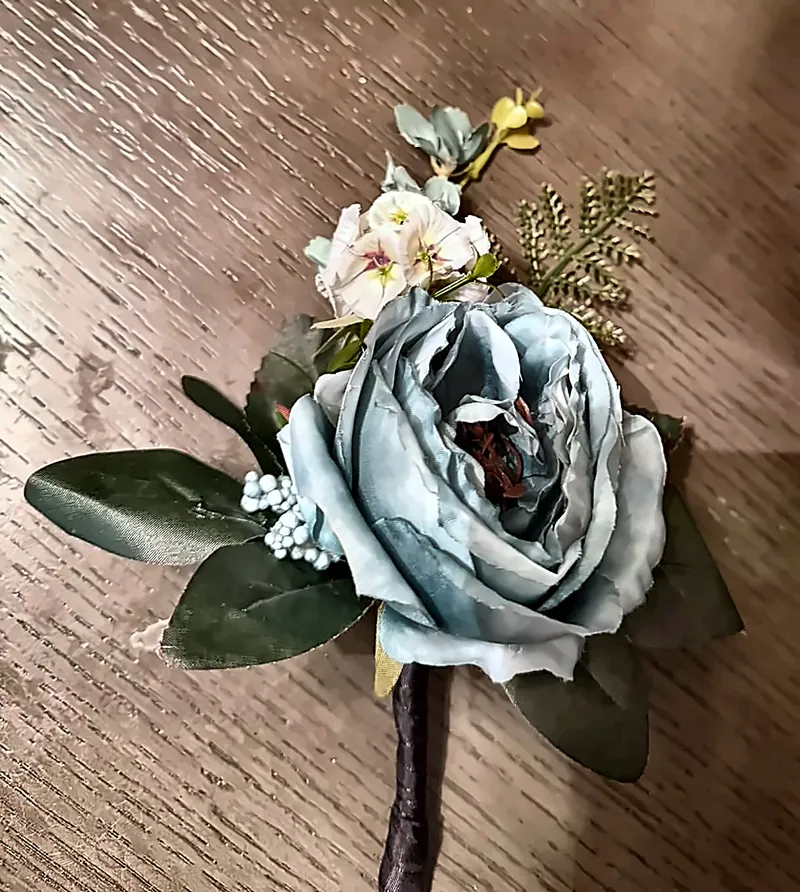 Boutonniere with a dusty blue rose, small blush flowers, fern, green leaves, and pale blue berries, with a ribbon-wrapped stem