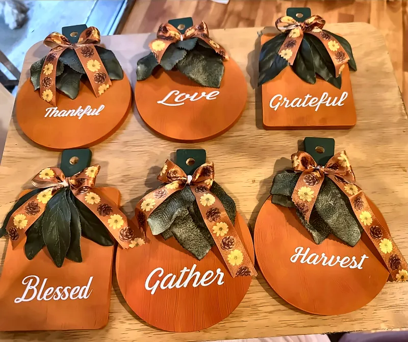 Six orange pumpkin-shaped wooden signs with white script words Thankful, Love, Grateful, Blessed, Gather and Harvest, each topped with green leaves and an autumn ribbon bow